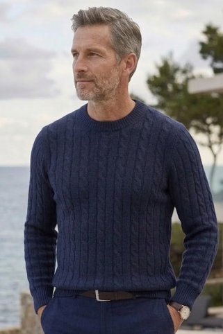 Muškarac u elegantnom pletenom džemperu stoji na terasi primorske vile s pogledom na ocean tijekom djelomice oblačnog dana.