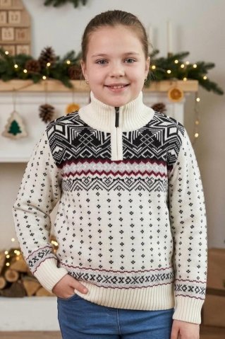 Usmívající se dívka v bílém pleteném svetru se zipem u krku a s tradičním norským vzorem stojí v interiéru zdobeném vánočními dekoracemi.