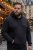 A robust man with a neatly trimmed beard stands on a city street in a black oversized hoodie, with one hand casually tucked into the front pocket, the other hanging relaxed by his side, while looking away to the left.