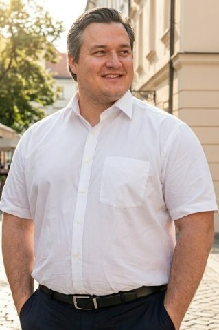 Elegantný korpulentný muž v bielej nadrozmernej košeli s krátkym rukávom stojí na slnečnej mestskej ulici s jednou rukou vo vrecku nohavíc.